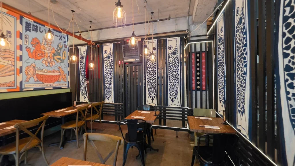 From left to right there is a wall booth with multiple tables along it. Then there are tables for two. The walls are decorated in artistic Japanese banners.